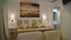 A double bed with floral pillows, a nightstand, a wall-mounted lamp, and a painting of a sunset over water. - Hotel Doña Lina (Seville)