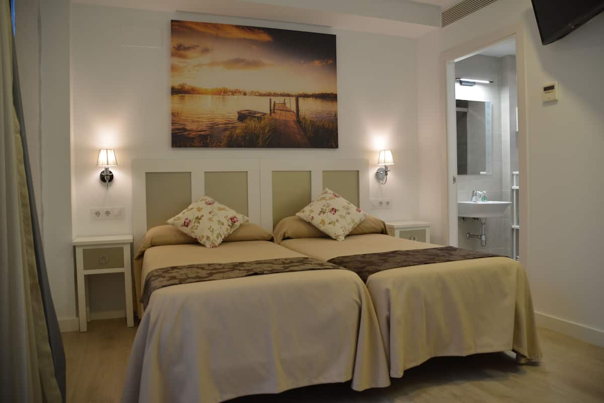 A double bed with floral pillows, a nightstand, a wall-mounted lamp, and a painting of a sunset over water.