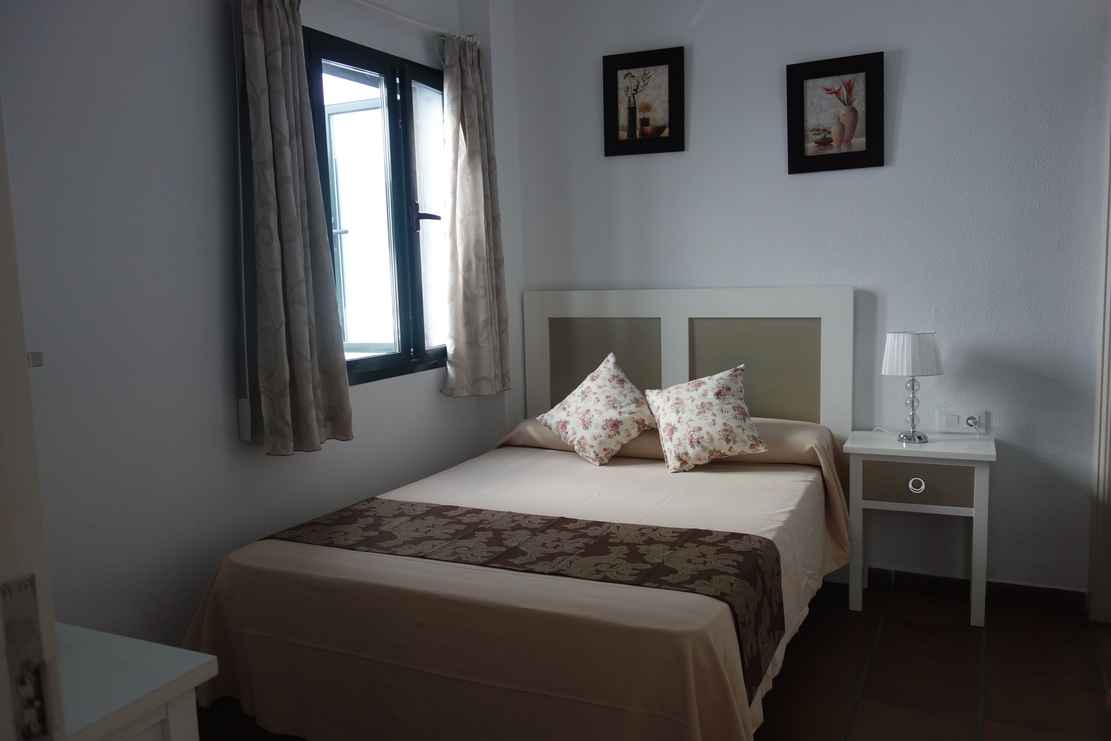 A bedroom with a bed, a nightstand, a lamp, and two wall-mounted pictures.