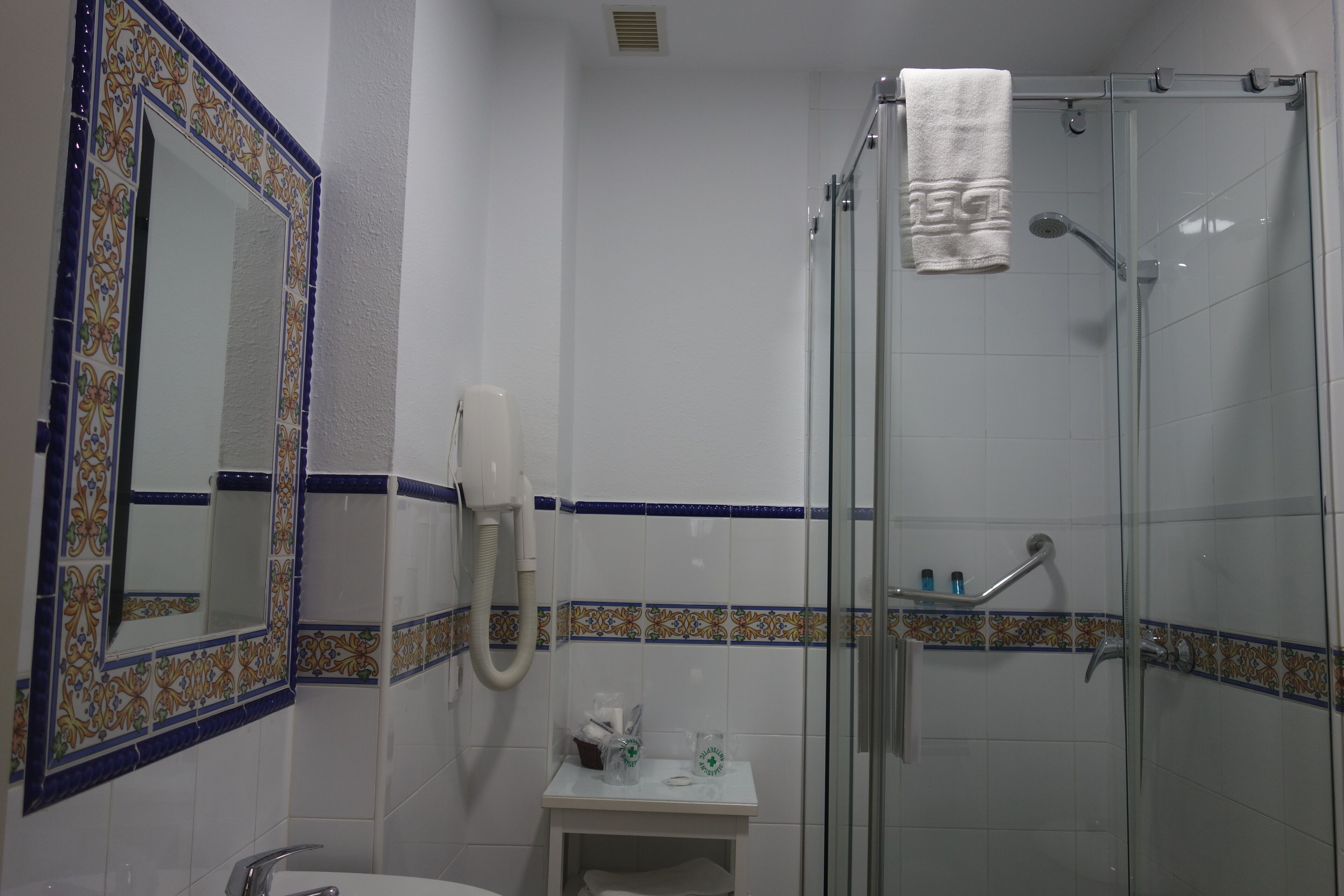 Un baño con ducha de mampara de vidrio, un lavamanos blanco, un espejo con marco decorativo y una toalla colgada en la puerta de la ducha.