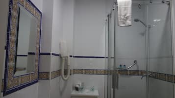 Un baño con ducha de mampara de vidrio, un lavamanos blanco, un espejo con marco decorativo y una toalla colgada en la puerta de la ducha.
