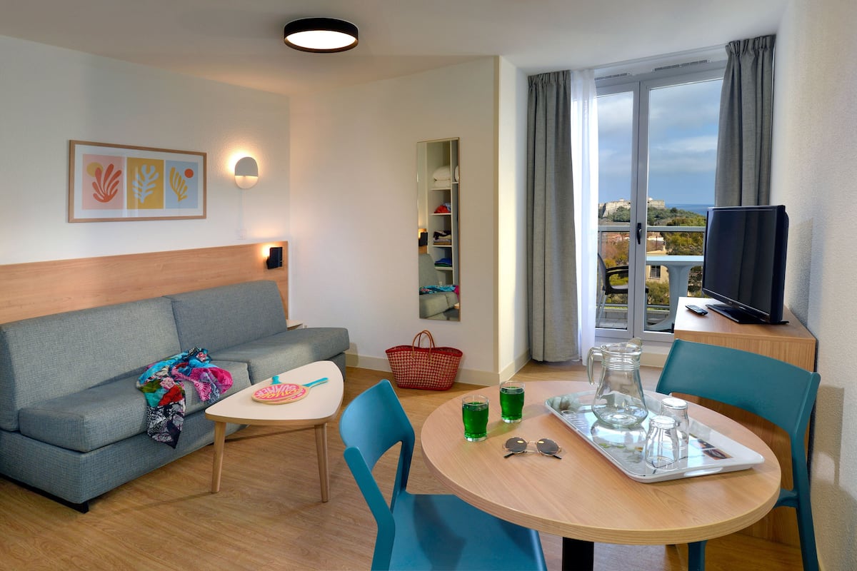 Quarto de hotel moderno com sofá, mesa de jantar com cadeiras, televisão e varanda com vista.