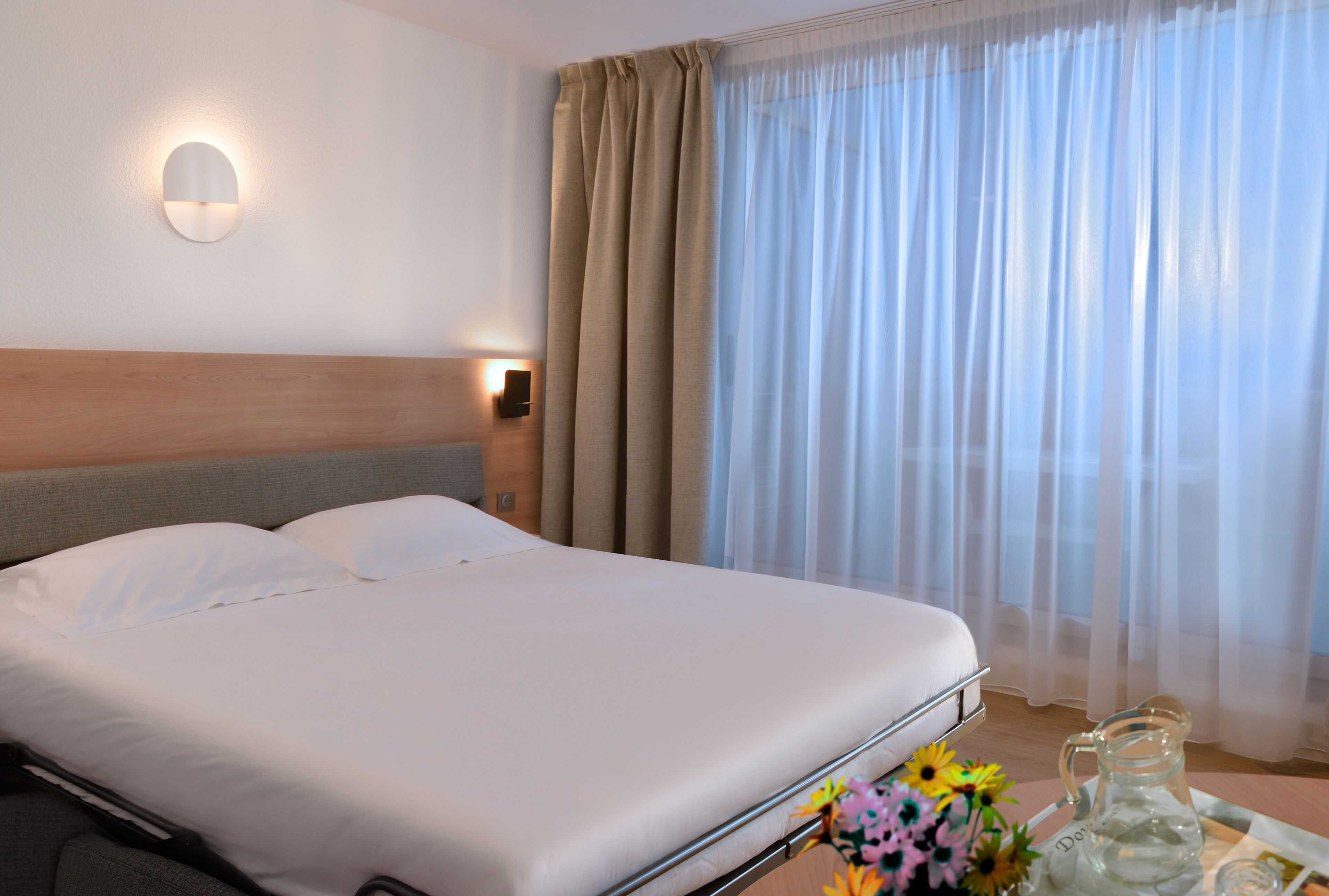 Une chambre d’hôtel avec un grand lit, une tête de lit, une table de chevet avec un pichet et des fleurs, et une fenêtre avec des rideaux légers.