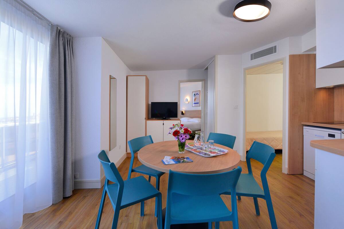 Quarto de hotel moderno com uma mesa de jantar redonda de madeira, cadeiras azuis, televisão de tela plana, pequena área de cozinha e uma grande janela com cortinas translúcidas.