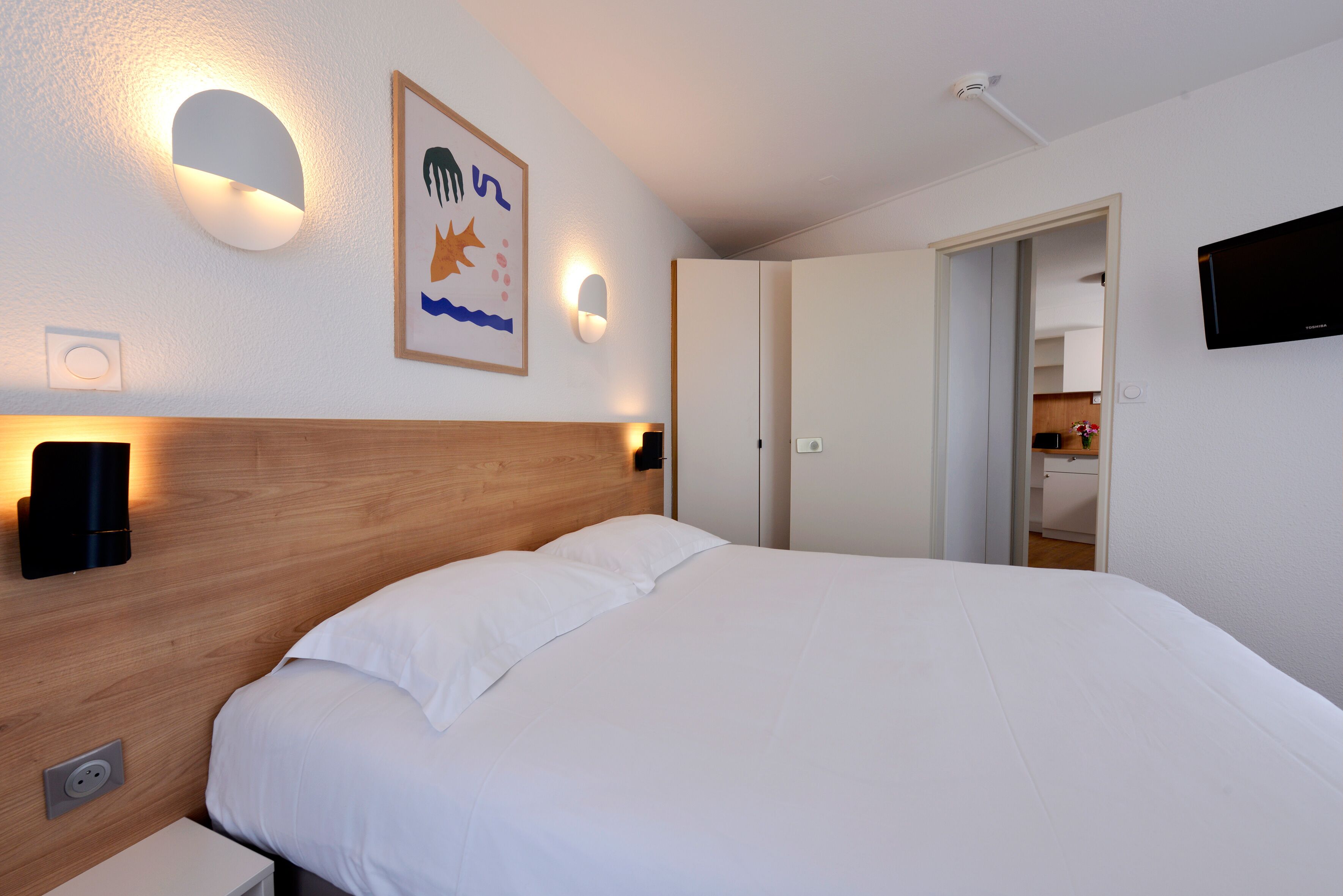 A bedroom with a wooden headboard, white bedding, wall-mounted light fixtures, and a framed picture.