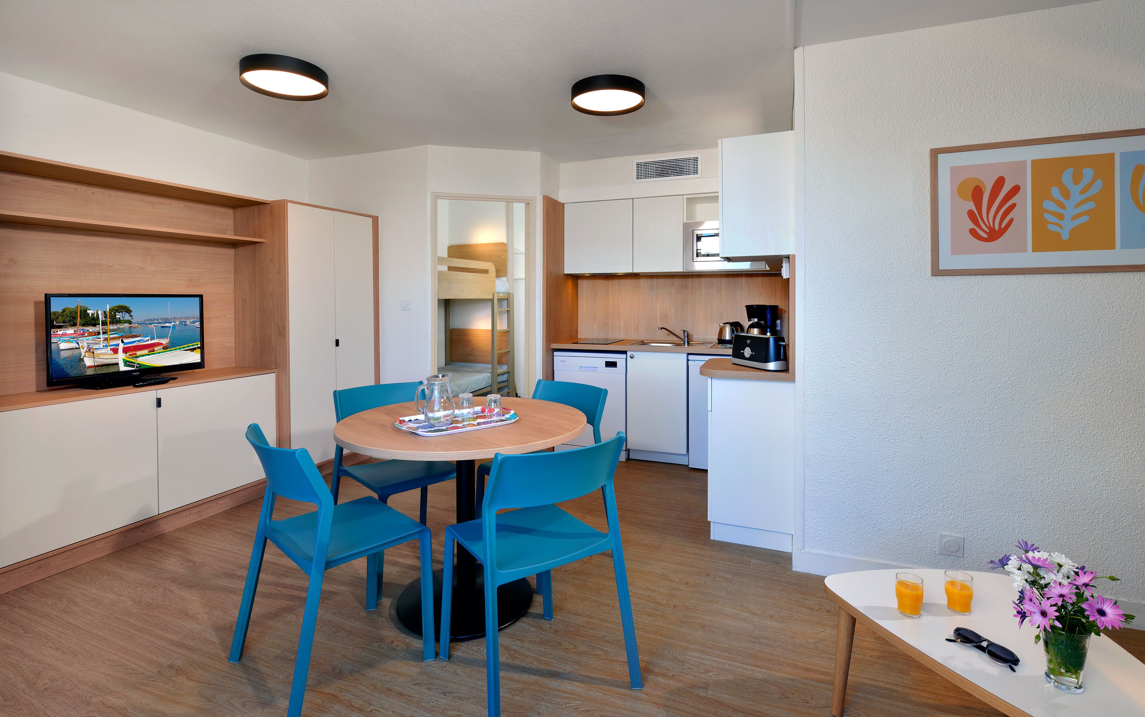 Una cocina compacta con electrodomésticos modernos, un comedor con sillas azules y un televisor mostrando una vista de puerto deportivo.