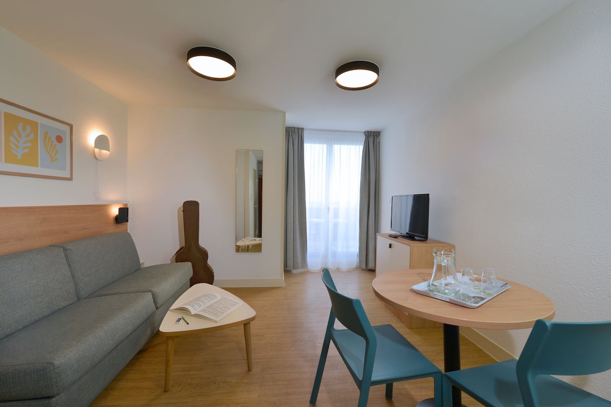 Quarto de hotel moderno com sofá, mesa de jantar com cadeiras, televisão e uma guitarra.
