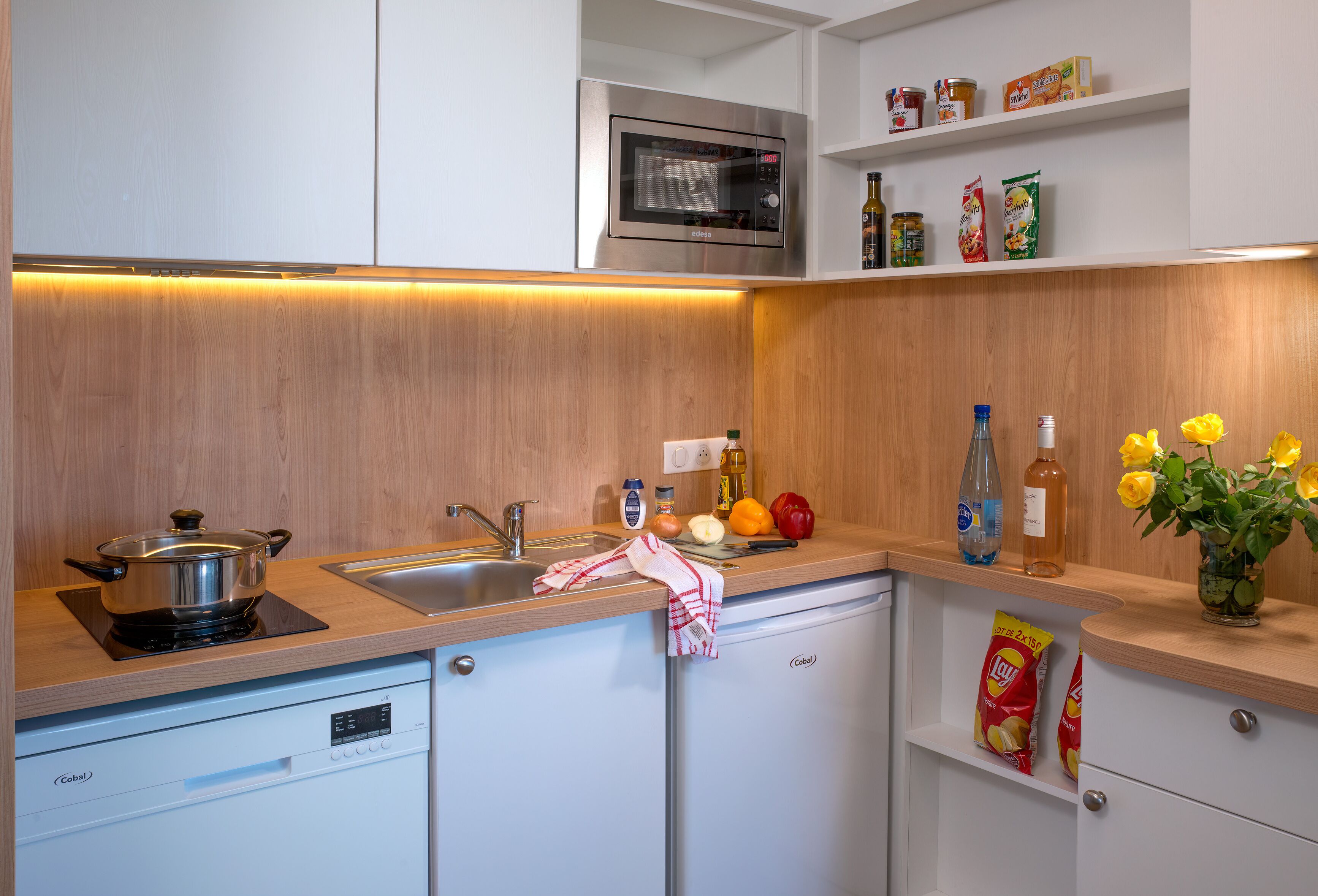 Une cuisine compacte équipée d’un four à micro-ondes, d’un évier et d’un petit réfrigérateur. On y trouve des placards, un plan de travail avec divers objets, et un pare-feu en bois.