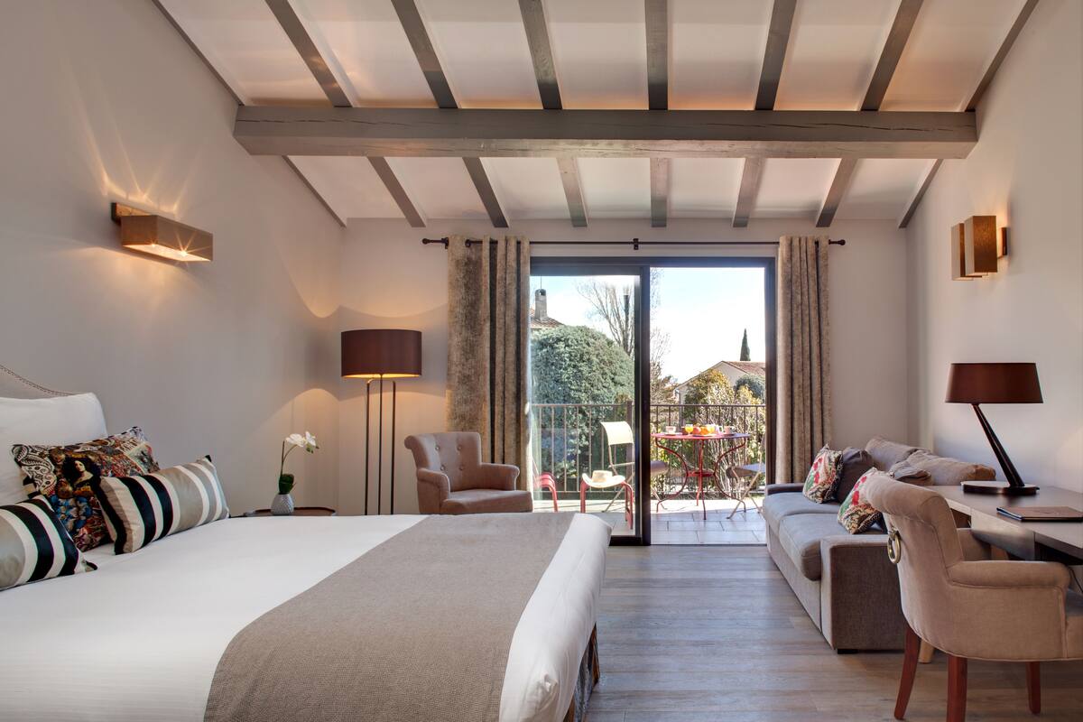 A modern hotel room with a large bed, a sofa, a desk, and a balcony with outdoor seating.