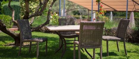 Un espacio para comer al aire libre con sillas de mimbre y una mesa, rodeado de follaje y una estructura de pérgola.