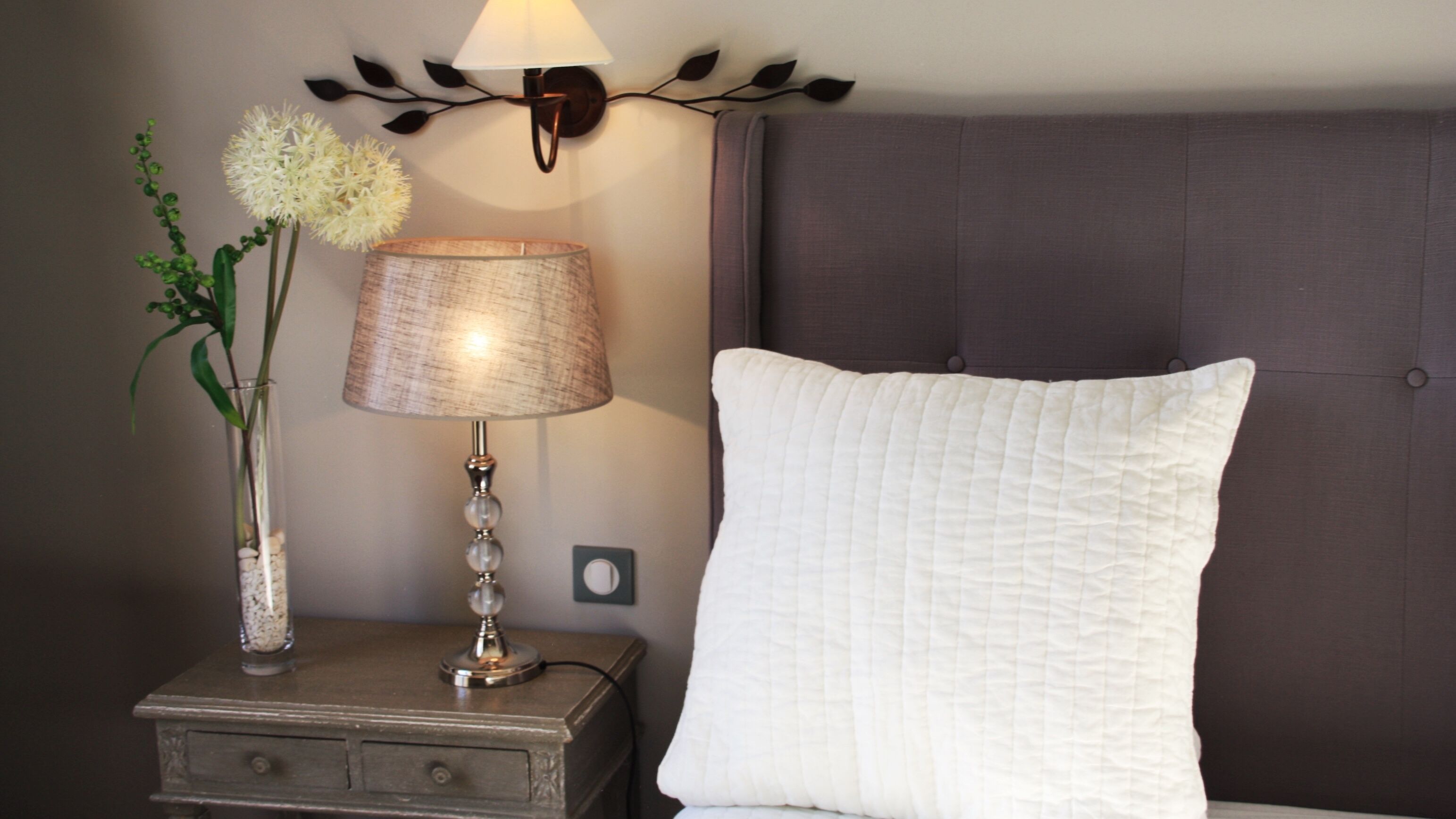 A neatly arranged bedroom with a bedside table, a lamp, a white pillow, and a decorative wall piece.