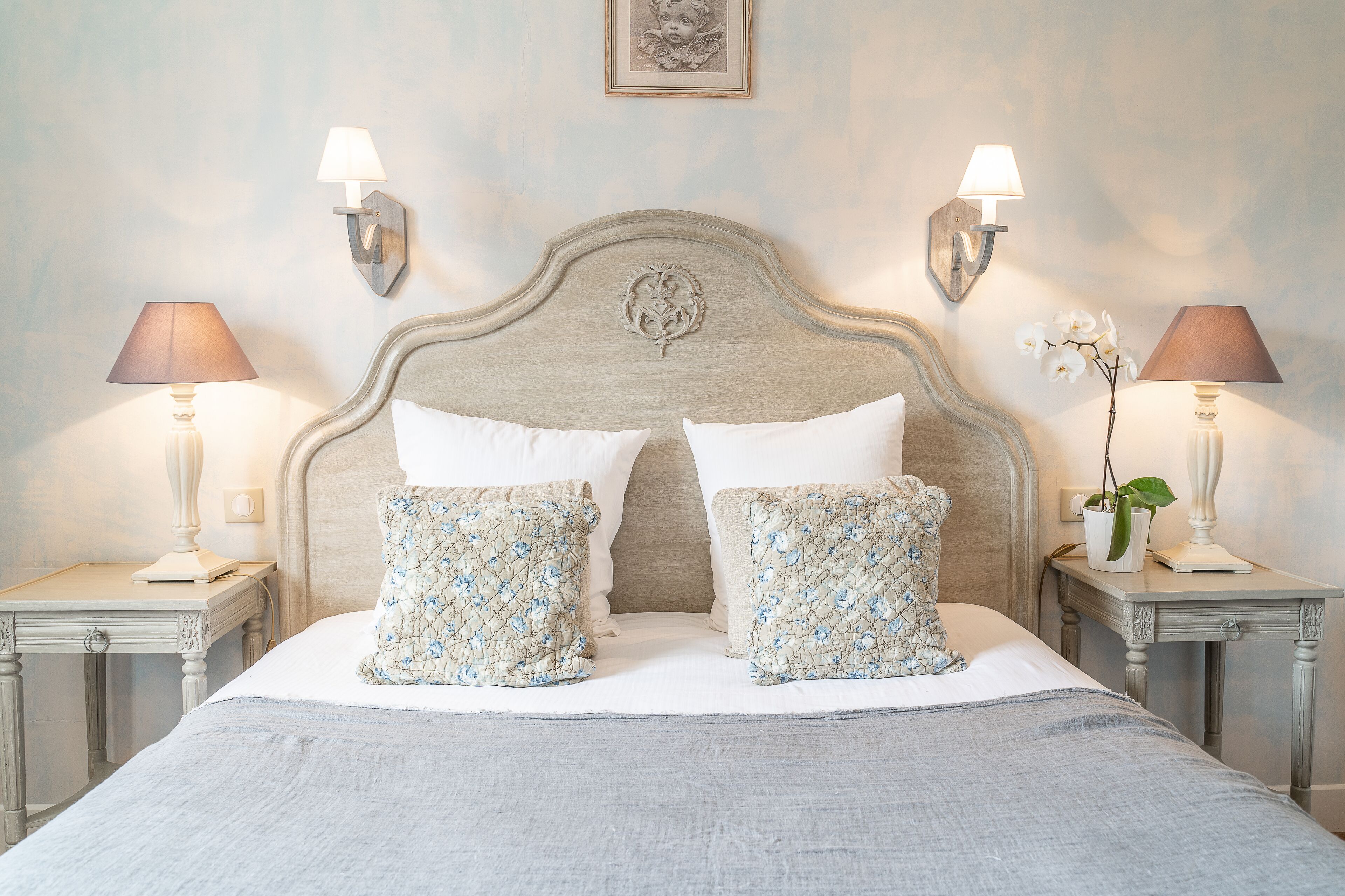 A neatly made bed with two pillows, two wall-mounted lamps, and a framed picture on the wall.