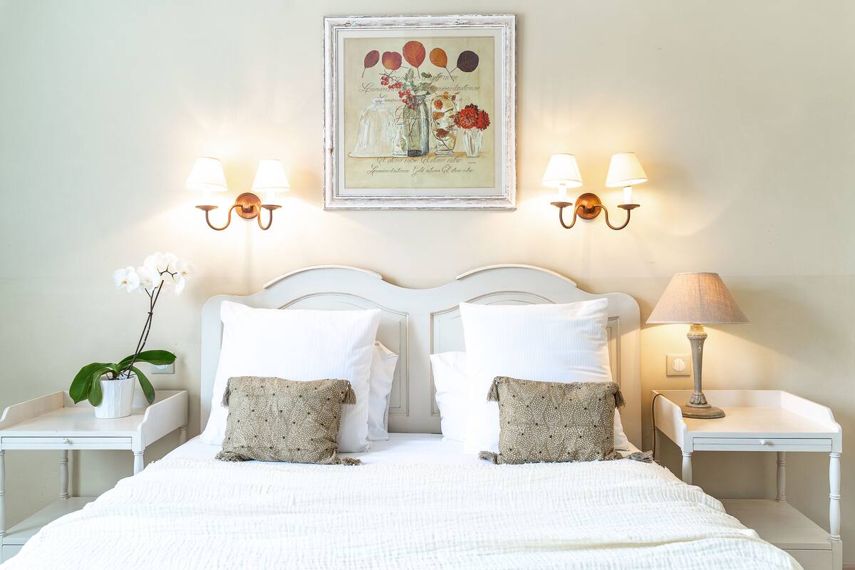 A neatly made bed with white linens and two decorative pillows, flanked by white nightstands with lamps and a framed artwork above.