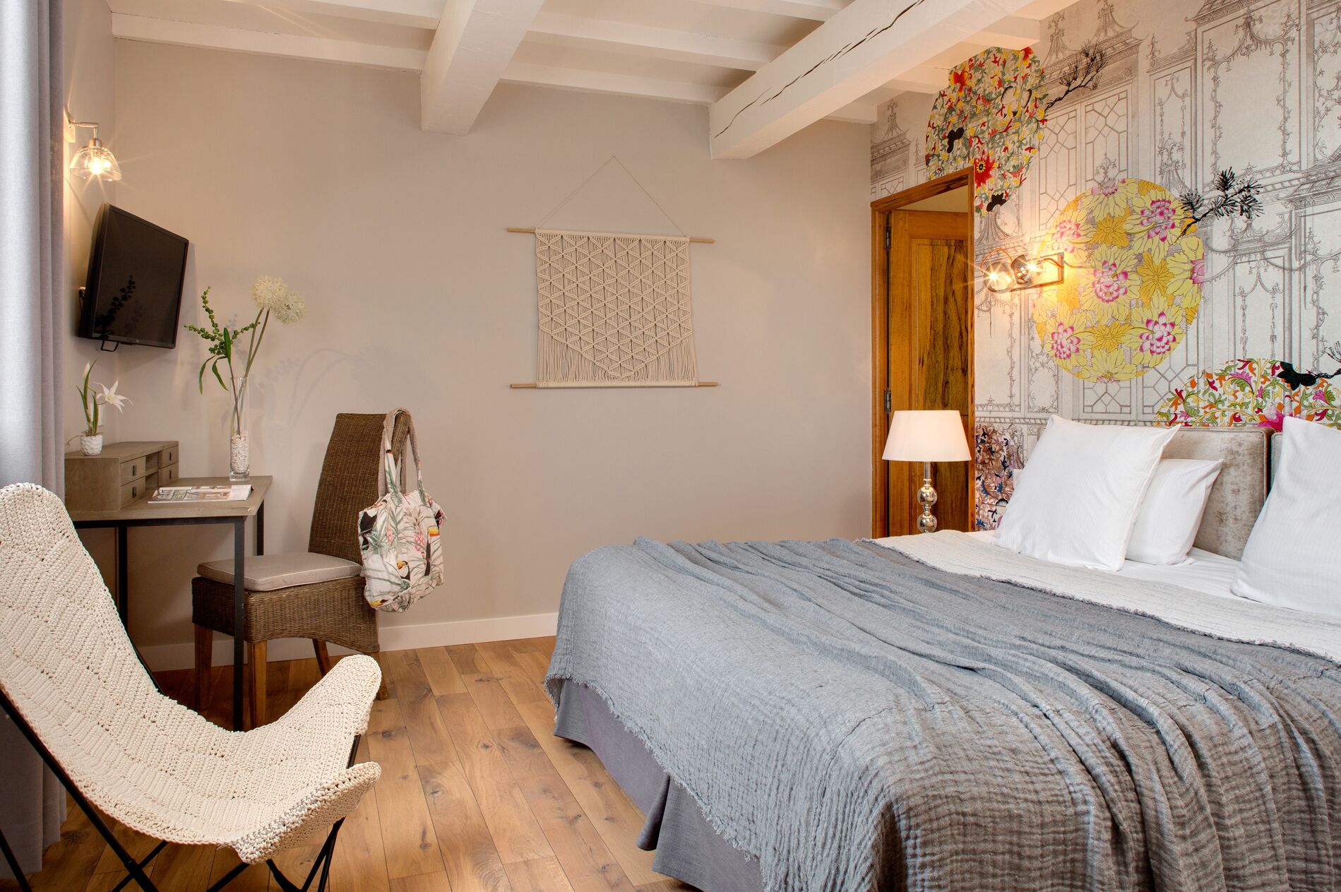 Ein Schlafzimmer mit einem Bett, einem Schreibtisch, einem Sessel, einem Fernseher und einer Wand mit Wandteppich mit Blumenmuster.