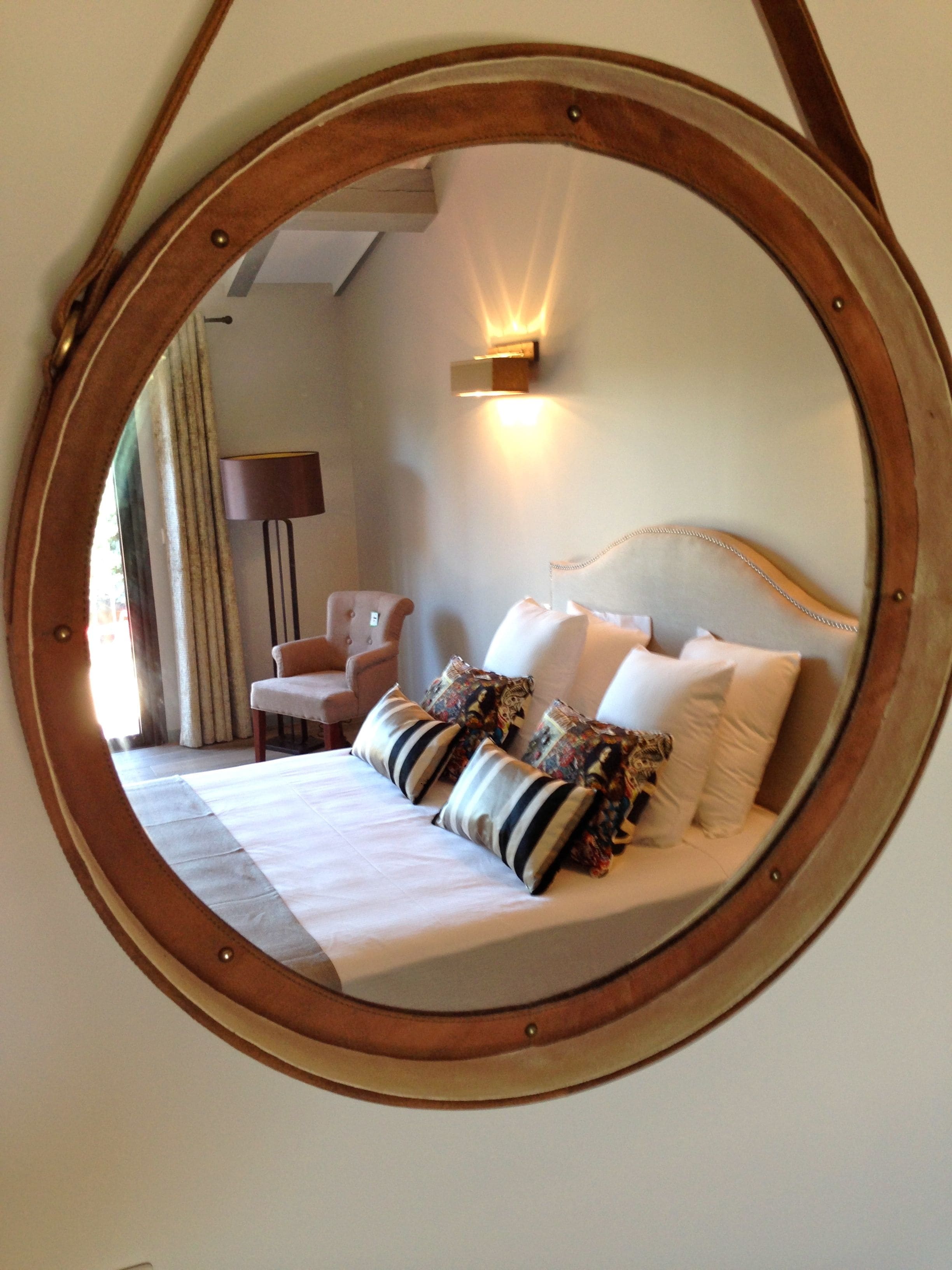 A circular mirror reflecting a bed with striped and patterned pillows, a bedside lamp, and a chair in a room with a neutral color scheme.