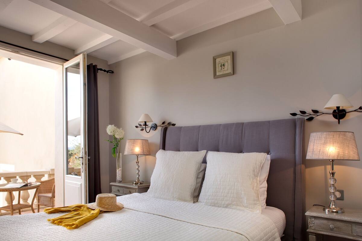 A bedroom with a bed, two bedside tables with lamps, a window with curtains, and a small framed picture on the wall.