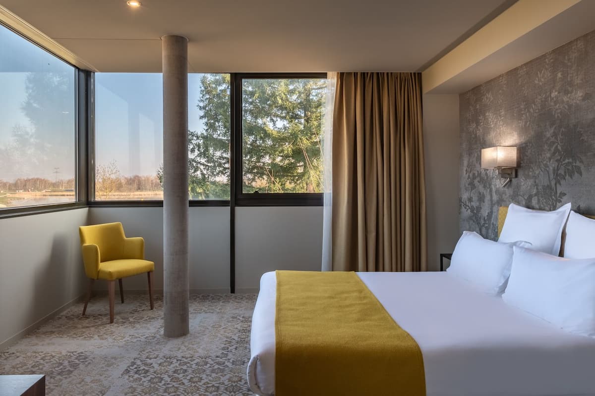 Une chambre d’hôtel avec un grand lit, une chaise jaune, une fenêtre donnant sur des arbres et un mur à texture.