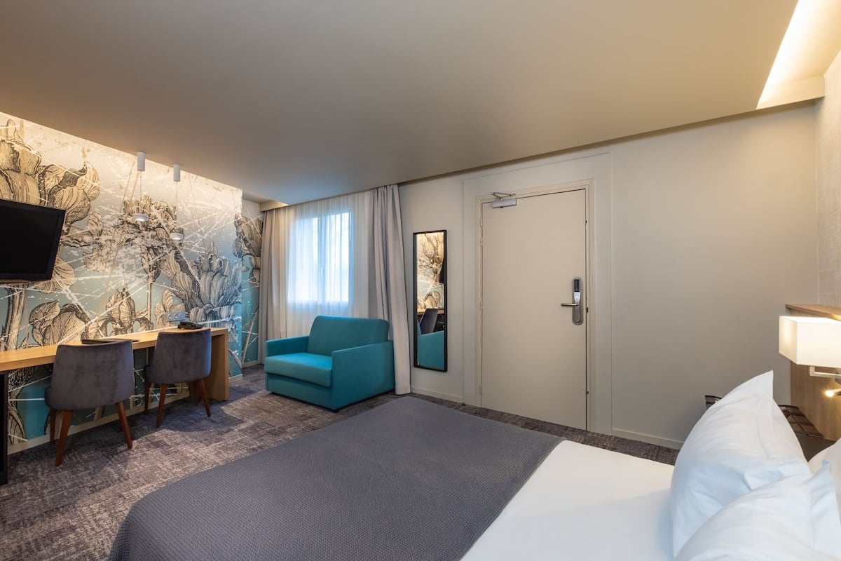 Une chambre d’hôtel comprenant un lit, un bureau avec deux chaises, un fauteuil de couleur turquoise, un grand miroir et une porte avec une serrure.