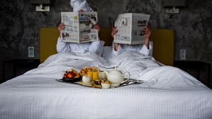 Twee personen in bed die kranten lezen, met ontbijtartikelen op een dienblad.