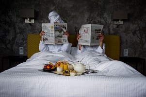 Dos personas en la cama leyendo periódicos, con una bandeja de desayuno.
