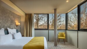 Een hotelkamer met een groot bed, een gele fauteuil en uitzicht op bomen door grote ramen.