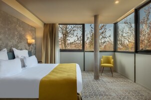 Habitación de hotel con una cama grande, un sillón amarillo y vistas a árboles a través de amplias ventanas.
