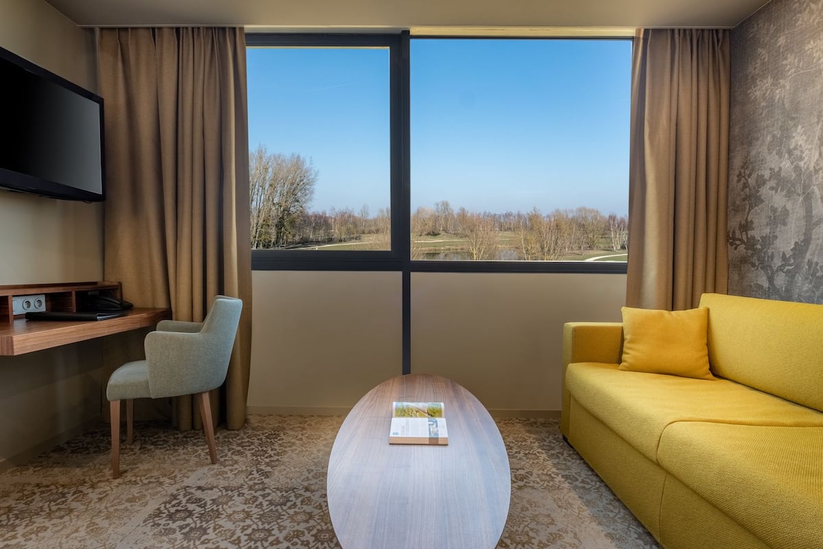 Une chambre d’hôtel équipée d’un bureau, d’une chaise, d’un canapé jaune, d’une table basse et d’une grande fenêtre donnant sur des arbres et une route.