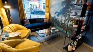 A modern living room with a dark blue sofa, yellow armchairs, a glass coffee table, and a bookshelf. A large window offers a view of the street outside. - ATH Hôtel Athénée (Cannes)
