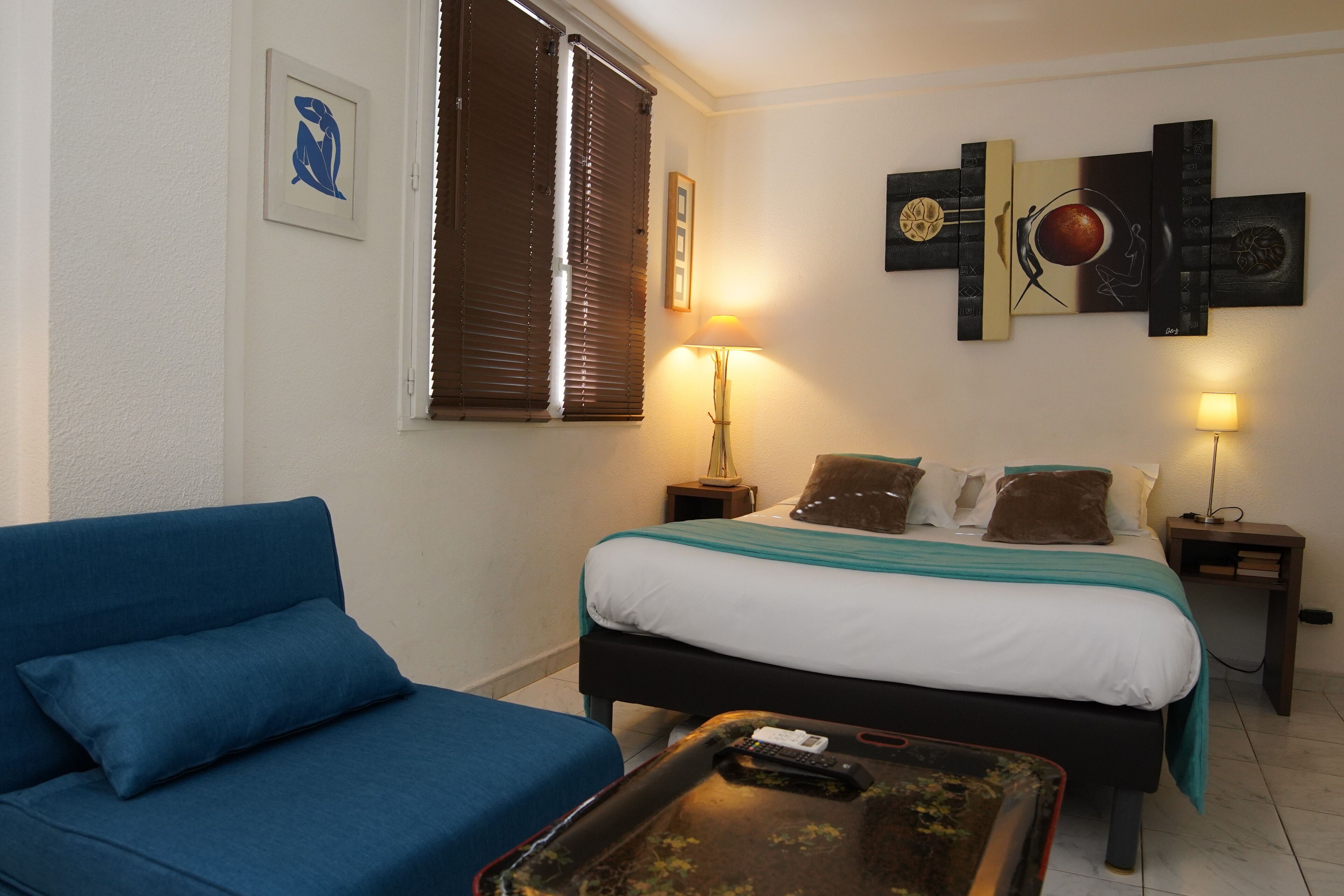 A bedroom with a bed, a blue sofa, a bedside table with a lamp, and a wall art piece.