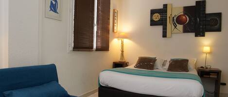 A bedroom with a bed, a blue sofa, a bedside table with a lamp, and a wall art piece.