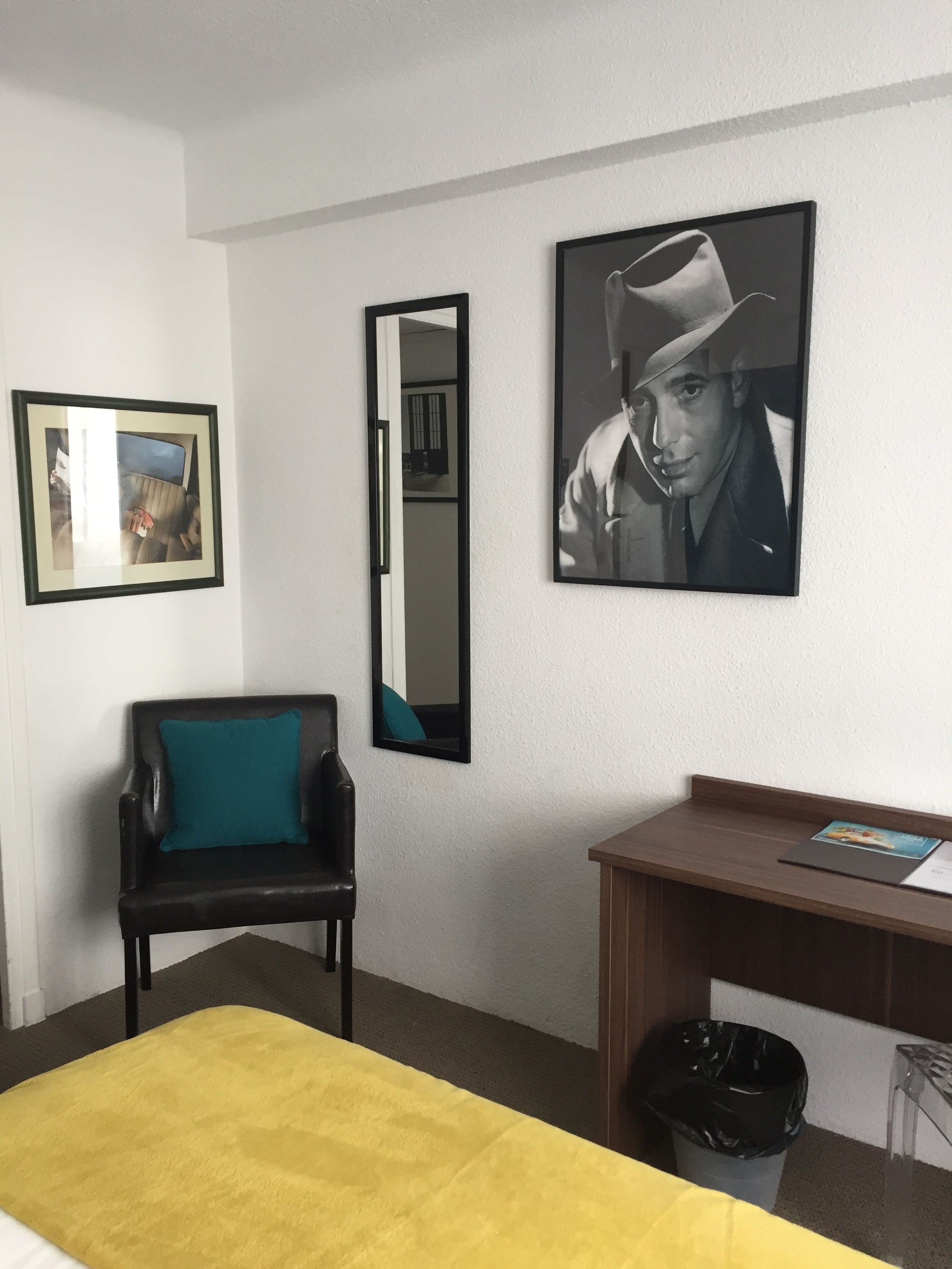 A hotel room with a bed, a desk, a chair, a trash bin, and two framed pictures on the wall.