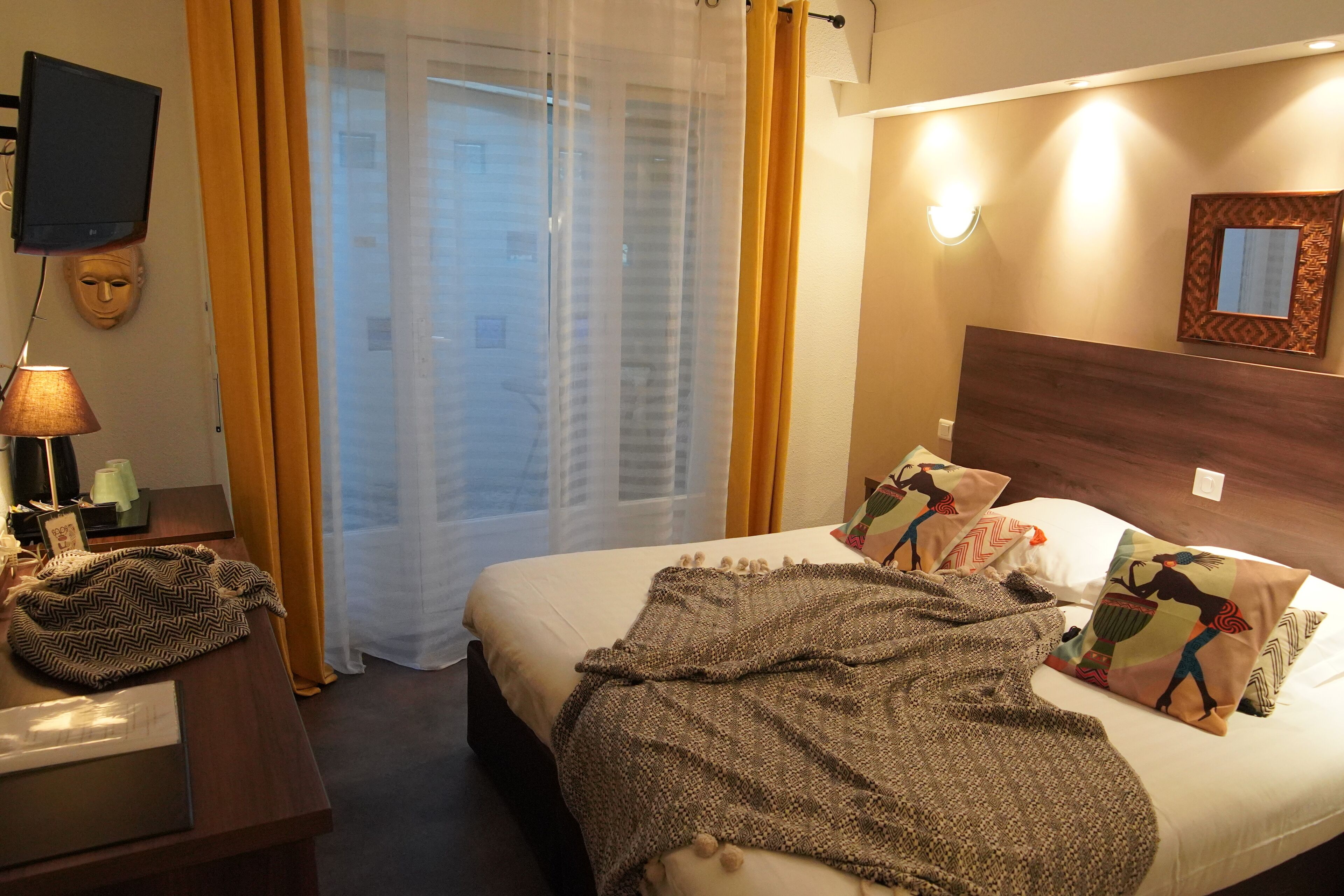 Hotellihuone, jossa on sänky, yöpöytä lampun kanssa, seinälle asennettu televisio ja ikkuna, jossa on verhot.