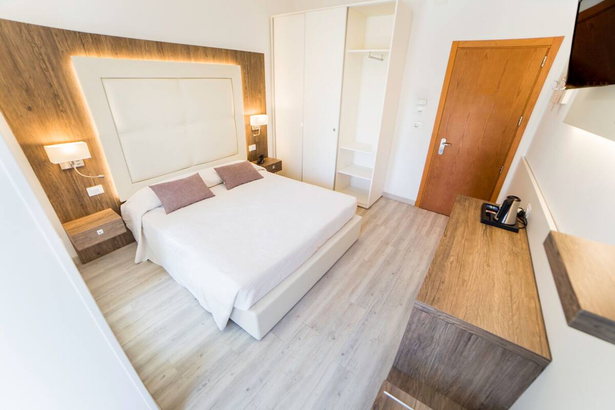 A modern hotel room with a large bed, bedside tables, a wardrobe, and a coffee machine.
