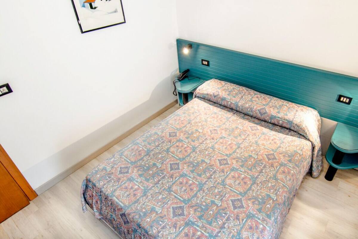 A small, neatly arranged bedroom with a patterned bedspread, a teal headboard, and a bedside table with a lamp.
