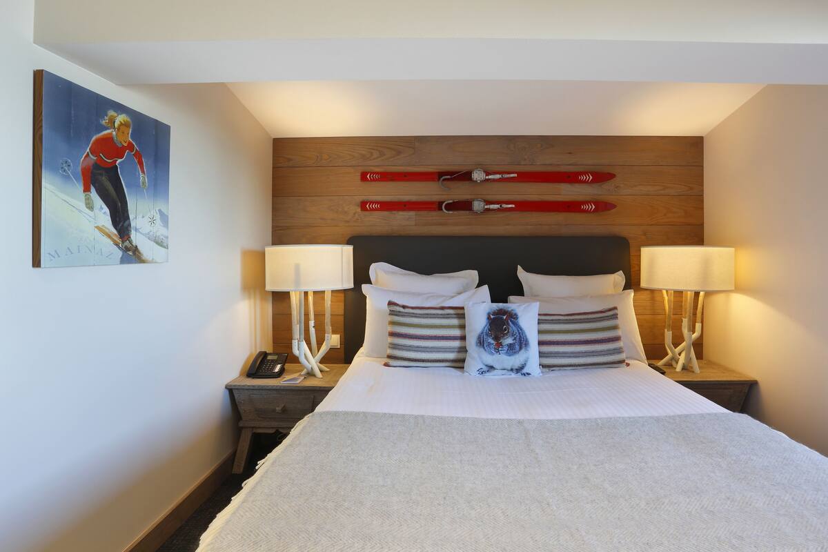 A neatly made bed with white linens, two bedside lamps, a wooden headboard, and a ski-themed artwork on the wall.