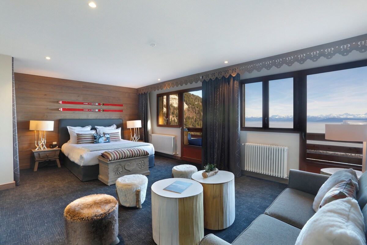 A modern hotel room with a large bed, two bedside tables, a sofa, and a view of mountains.