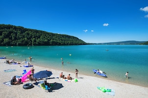 Uma praia com águas turquesa cristalinas, pessoas nadando e tomando sol, e uma colina arborizada ao fundo.