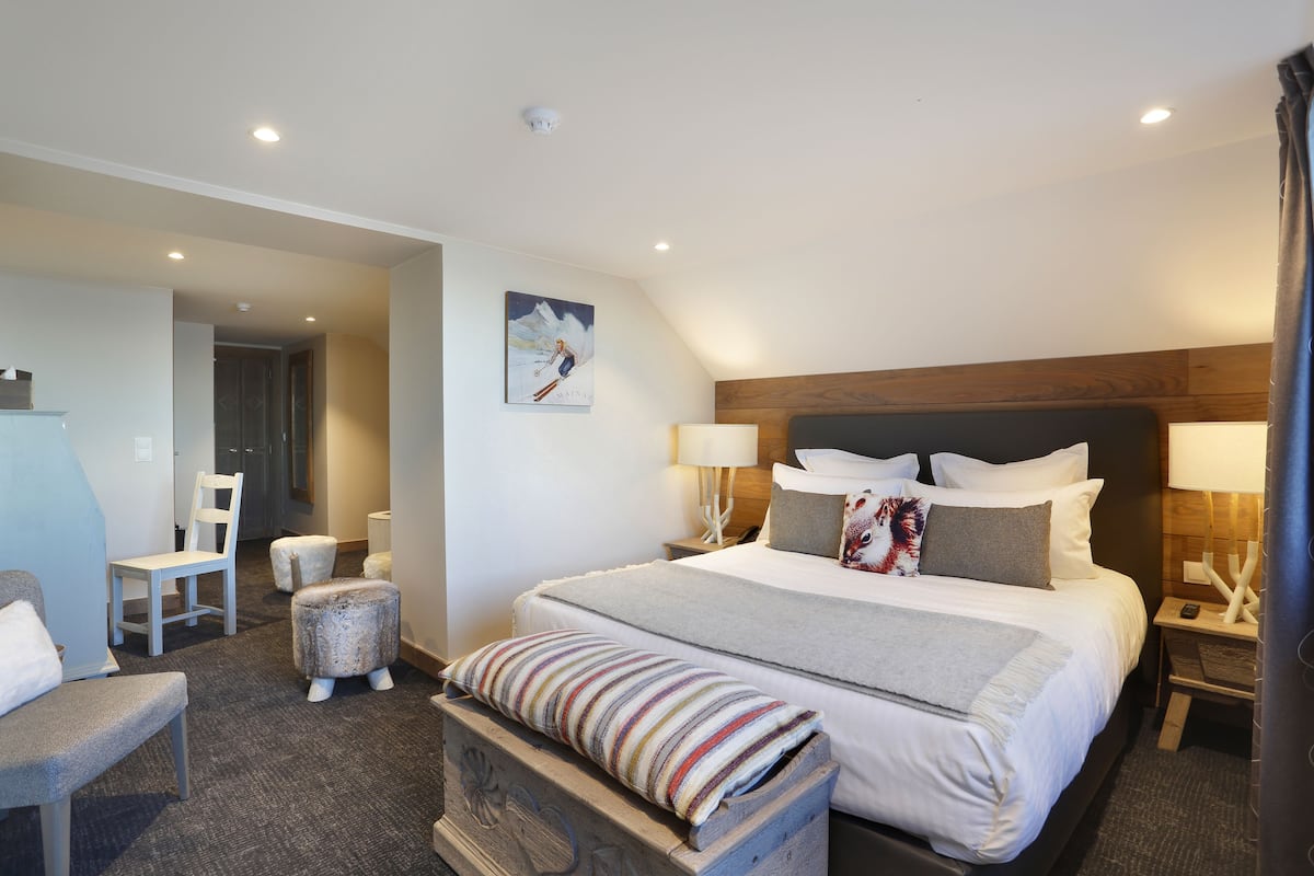 A modern hotel room with a large bed, a wooden headboard, two bedside tables with lamps, a chair, and a small ottoman.
