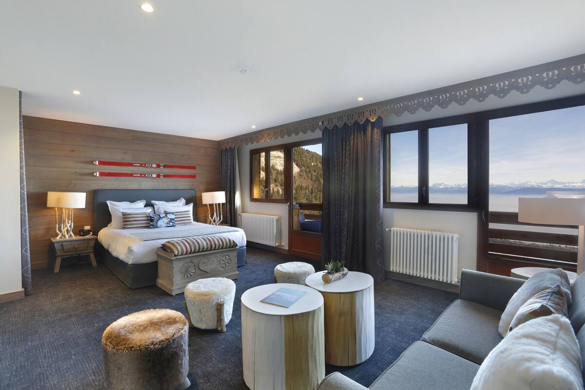 A modern hotel room with a large bed, two bedside tables, a sofa, and a view of mountains.