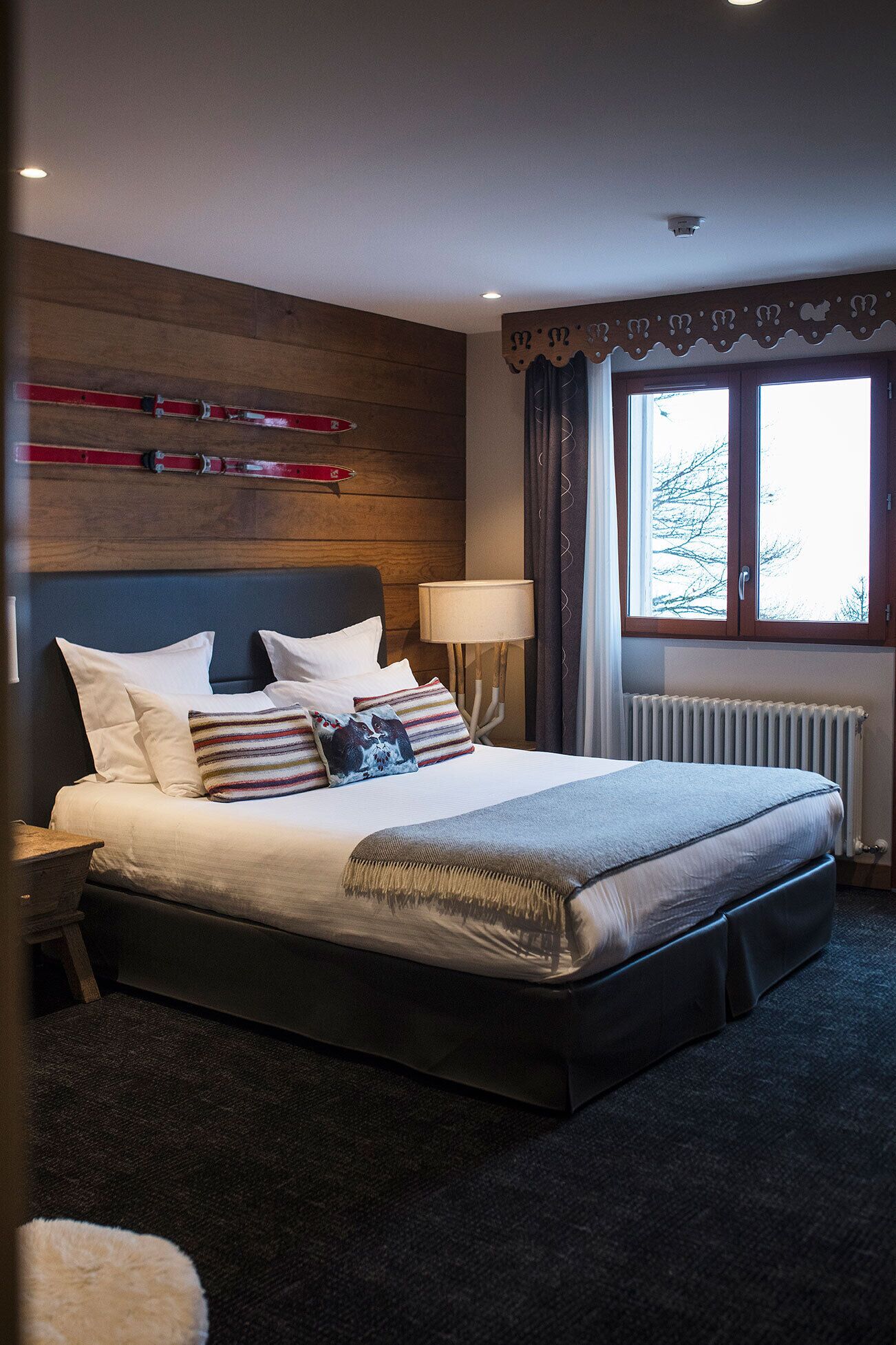 Une chambre d’hôtel avec un grand lit, une tête de lit en bois, une fenêtre avec des rideaux, un radiateur et une lampe.