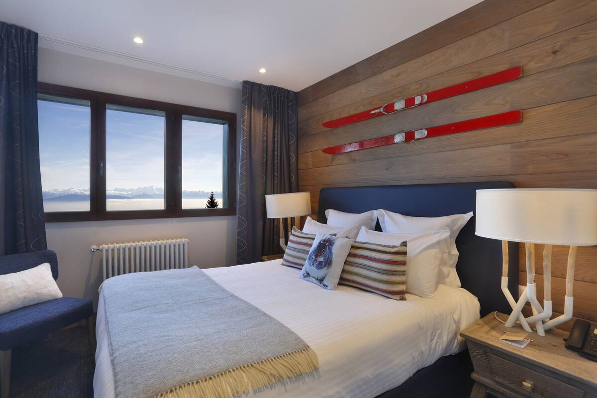 A modern hotel room with a large bed, two bedside tables, a view of the mountains, and wooden paneling on the walls.