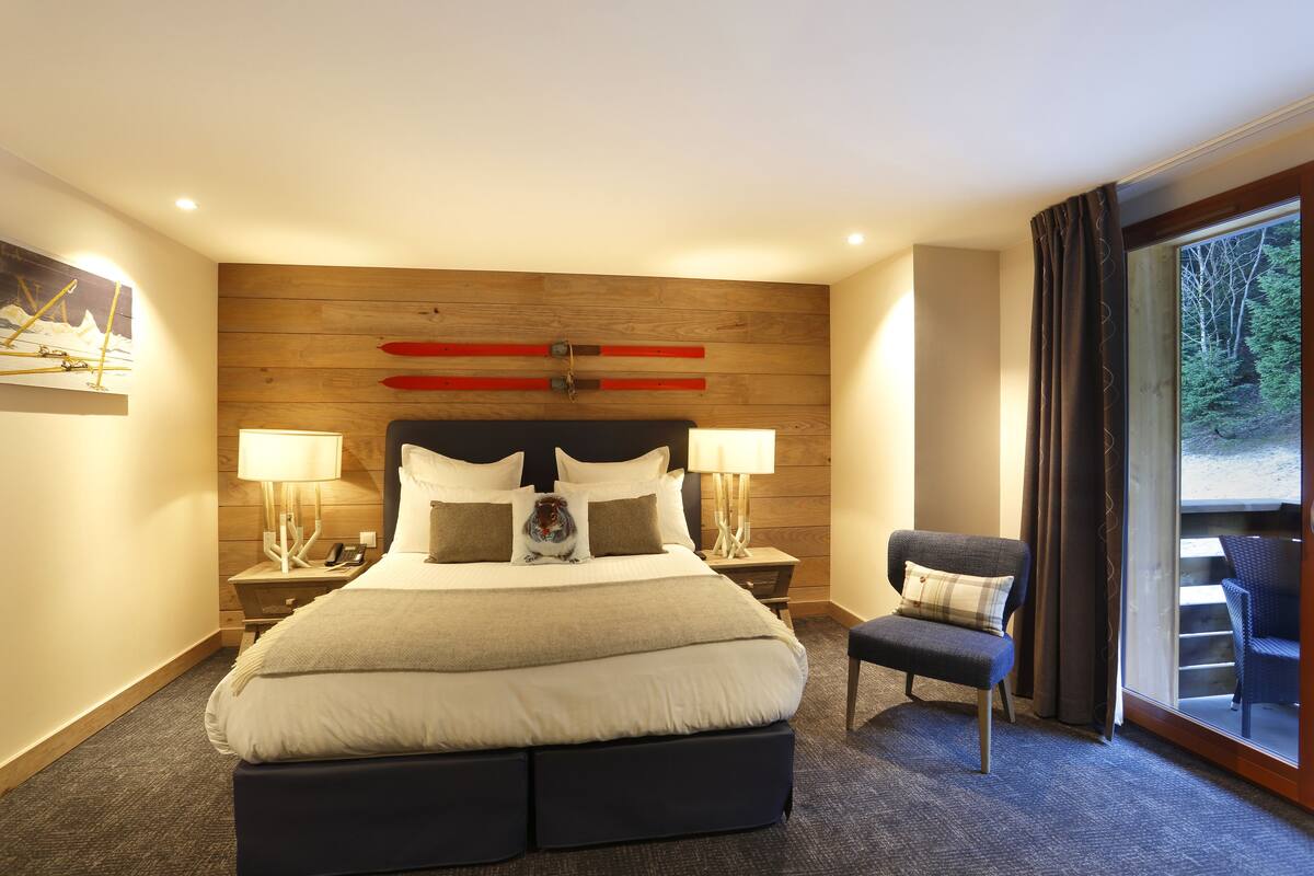 A hotel room with a large bed, two bedside tables with lamps, a chair, and a view of the outdoors through a sliding door.