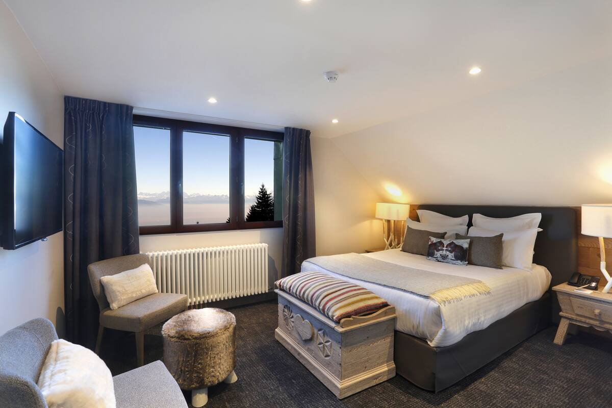 A modern hotel room with a large bed, a seating area with a chair and ottoman, a television mounted on the wall, and a window with a view of the mountains.