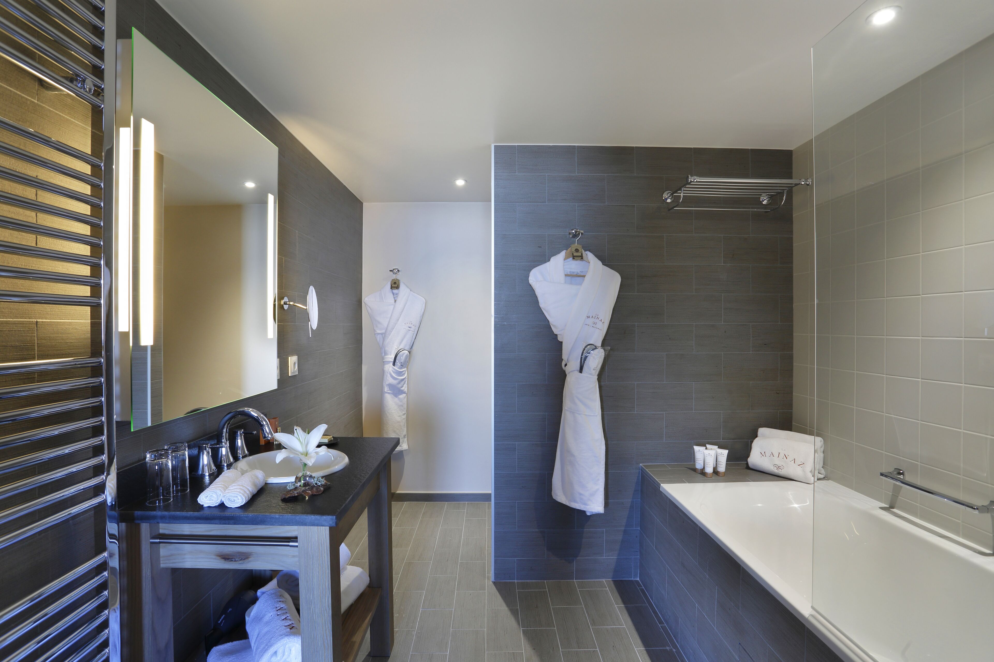 A modern bathroom with a bathtub, a vanity with a mirror, and two white robes hanging on hooks.