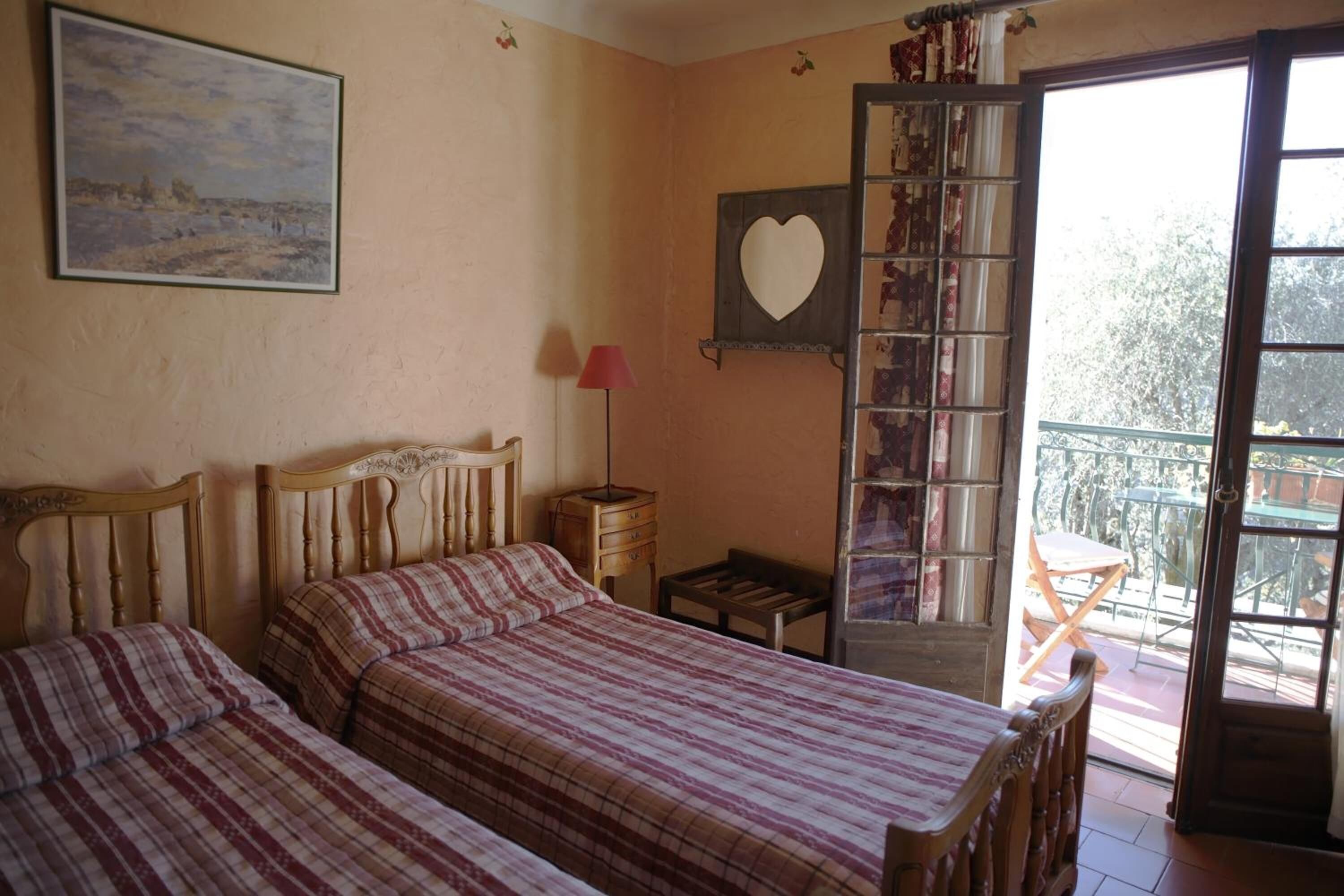 A bedroom with two beds, a wooden headboard, a bedside table with a lamp, a heart-shaped mirror, and a view of a balcony with outdoor furniture.
