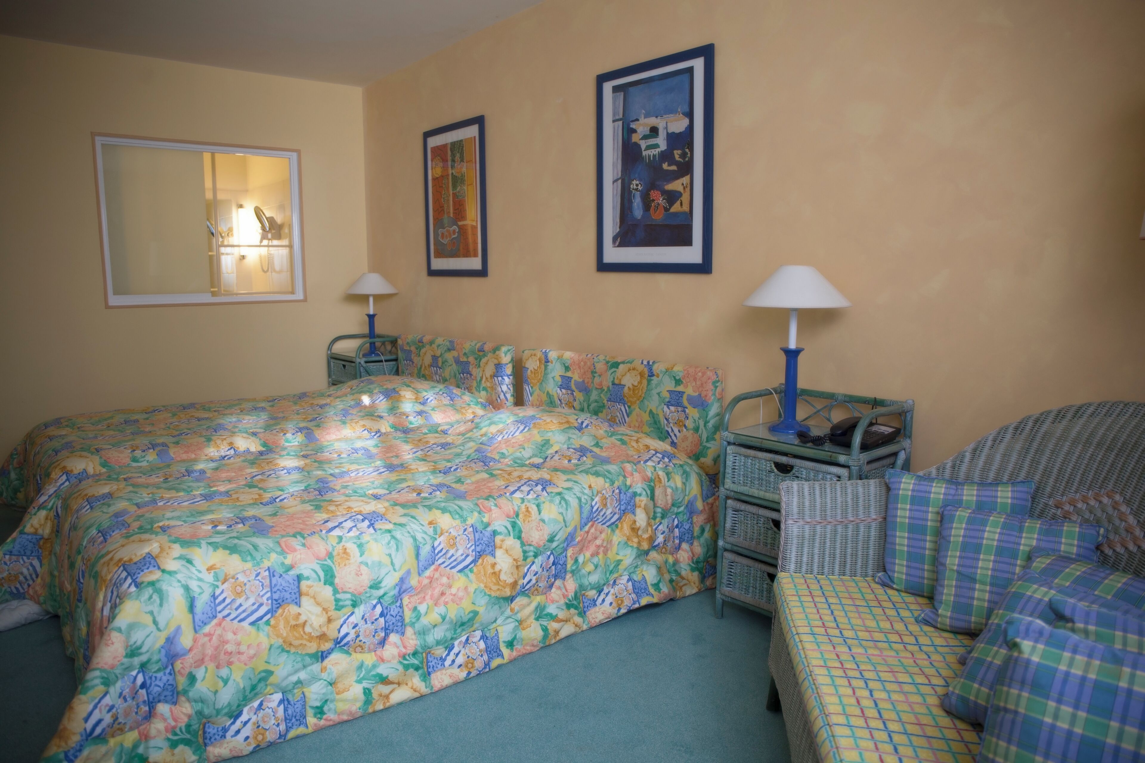 A bedroom with a floral bedspread, a large mirror, two wall-mounted lamps, and a wicker side table.