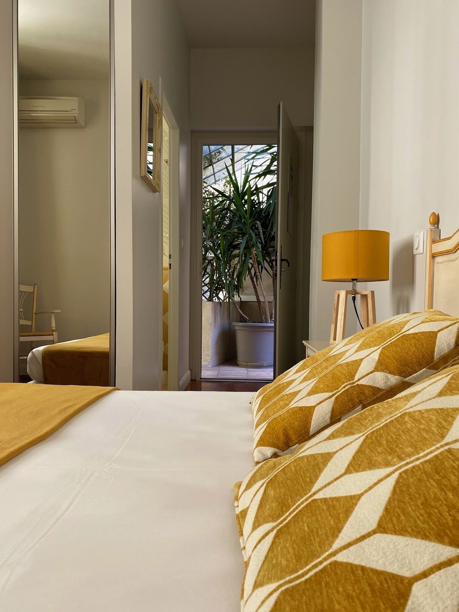 A hotel room with a bed, a bedside lamp, a chair, a potted plant, and an air conditioning unit.