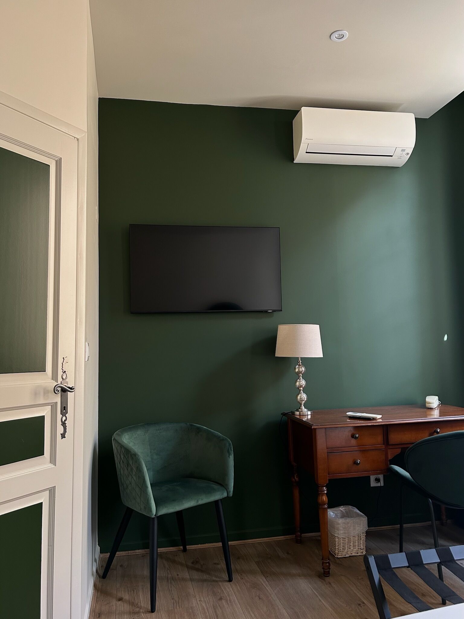 A room with a green wall, a flat-screen TV mounted above a wooden desk, a green chair, a lamp, and a white air conditioning unit.