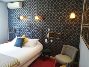 Habitación de hotel con papel tapiz estampado, una cama con ropa de cama blanca y azul, un taburete pequeño verde y un sillón gris.