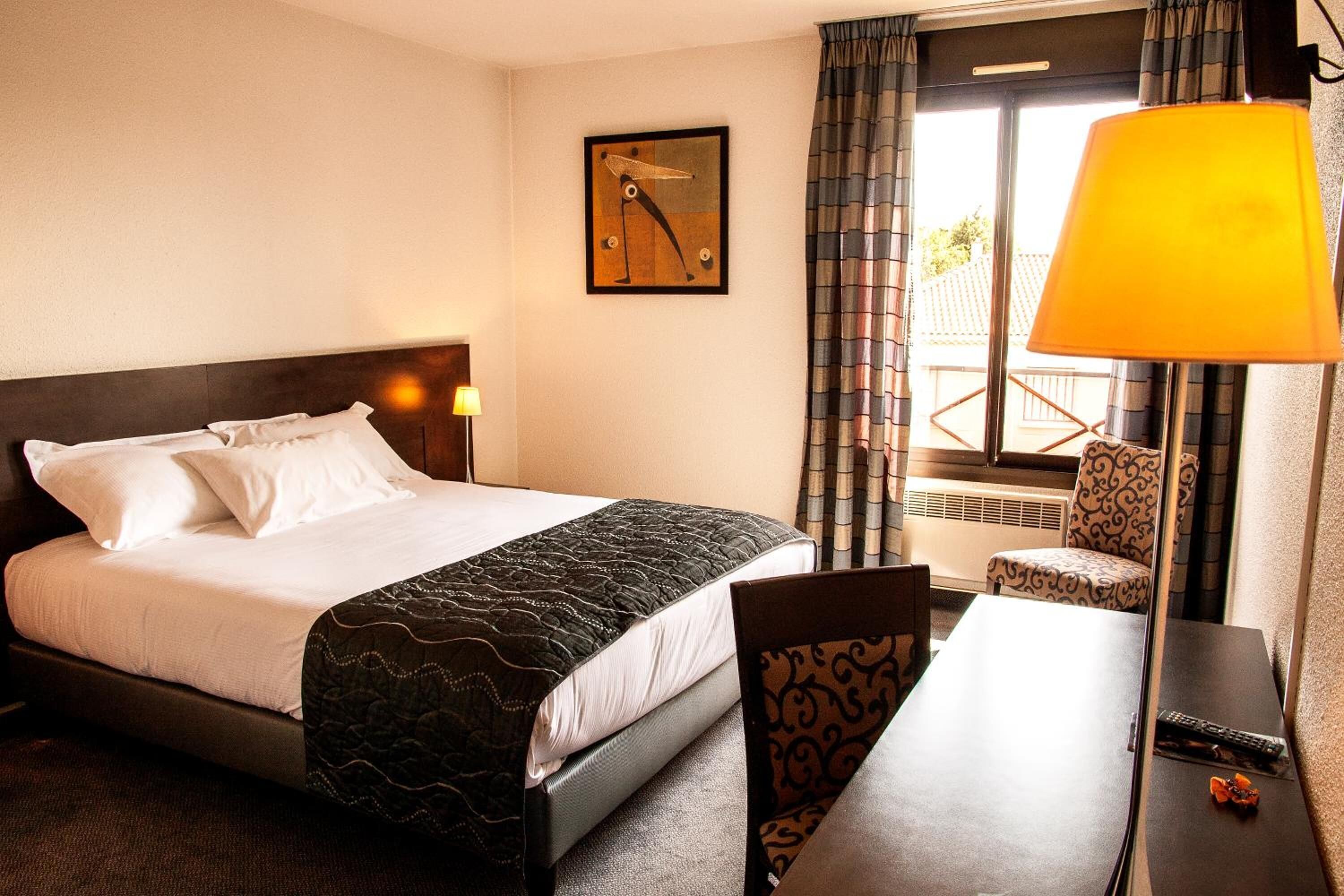 Ein Hotelzimmer mit einem Bett, einem Schreibtisch, einem Stuhl, einer Lampe, einem Fenster mit Vorhängen und einem Wandbild.
